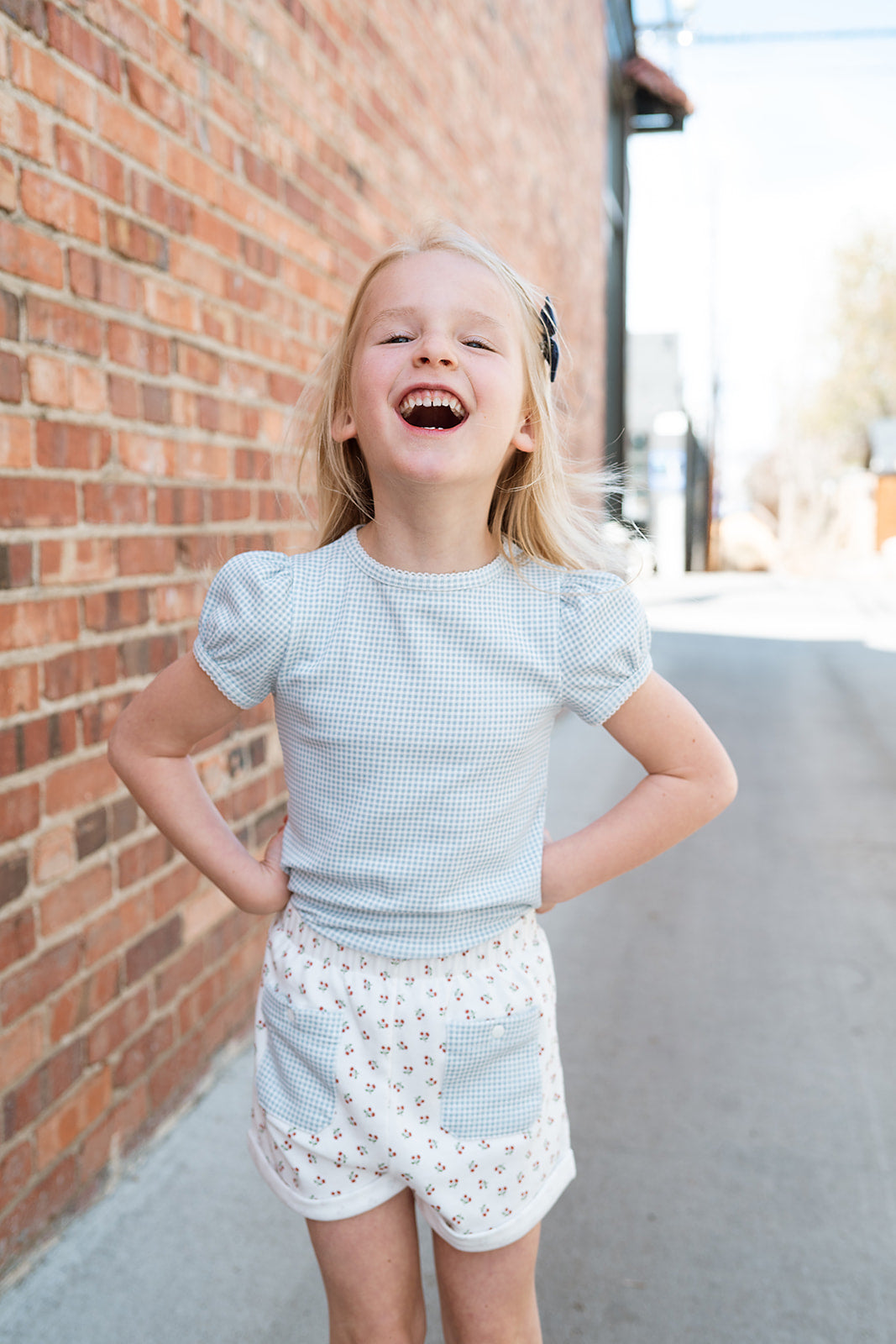 Little girl wearing a short sleeve light blue gingham tee and cherry print shorts made of high quality ethically produced organic pima cotton.