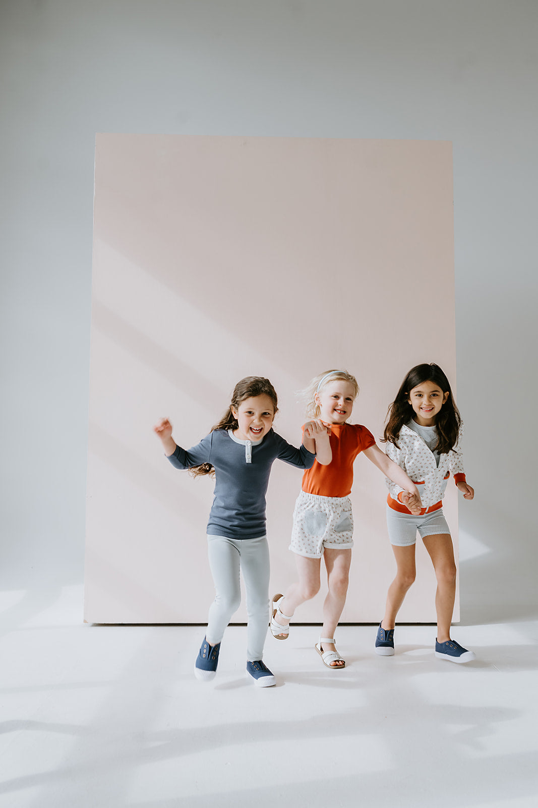 three kids running in front of a pink wall holding hands wearing organic pima cotton kids clothing with nostanglic styles and playful prints made for play.
