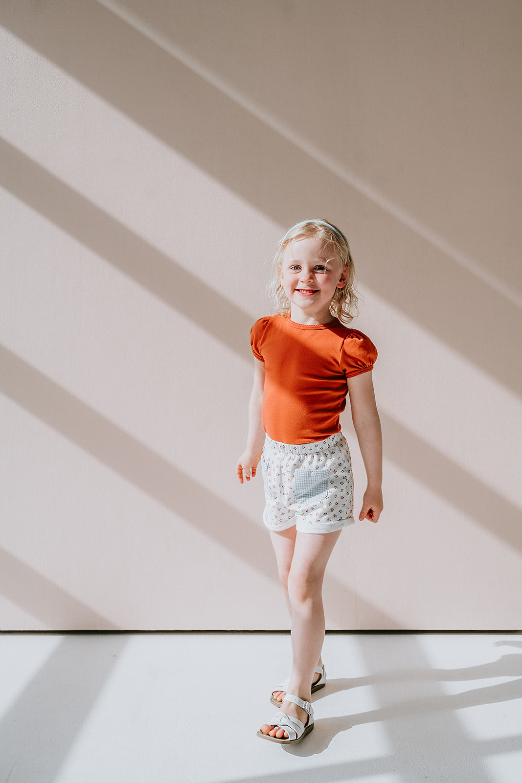 Little girl standing in front of pink wall in photography studio with light streaming in. She is wearing a red organic pima cotton girls tshirt and cherry print shorts with blue gingham pockets.