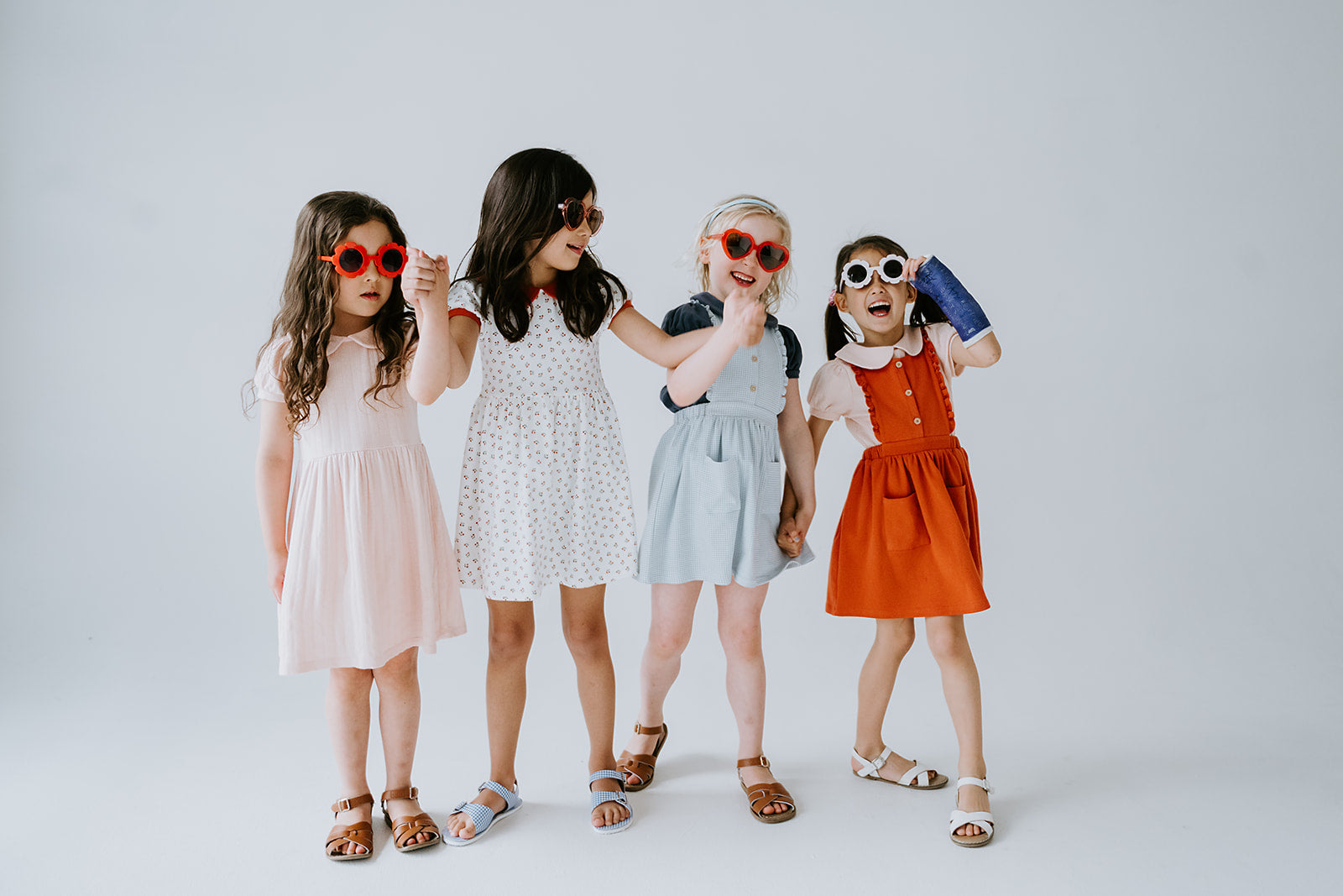 Four young girls in colorful dresses and sunglasses posing together on a white background