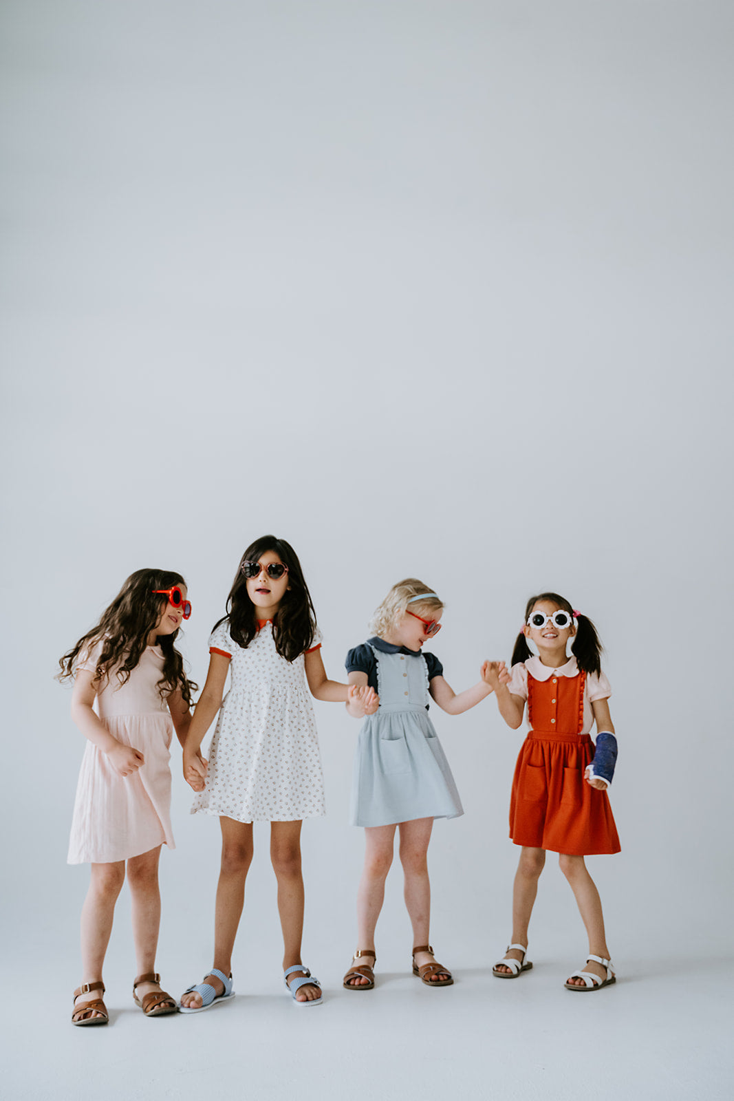 Four little girls standing in a white photography studio holding hands wearing flower and heart sunglasses. They are all wearing organic pima cotton girls dresses.
