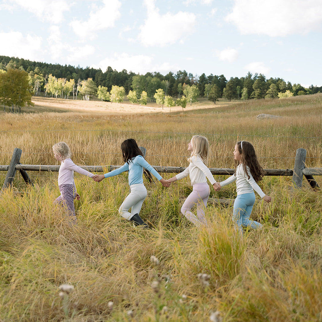 Four little girls holding hands walking along a wooden fence wearing organic pima cotton kids clothing.