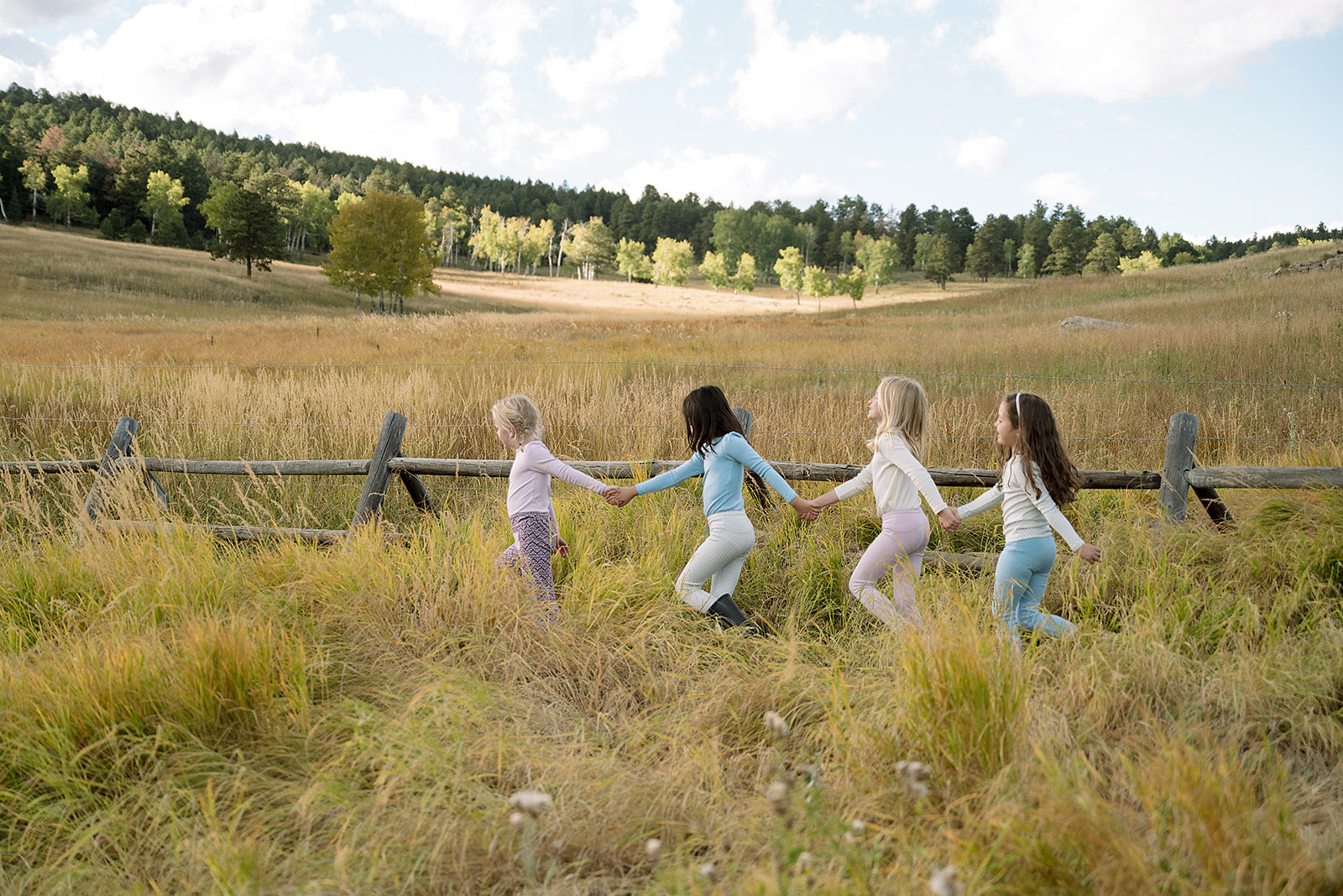 Four little girls holding hands walking along a wooden fence wearing organic pima cotton kids clothing.