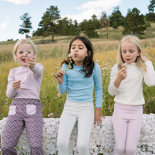 Three young girls blowing dandelion seeds in a field with trees in the background. They are all wearing organic pima cotton childrens clothing styles. A mix of light blue, cream pointelle, and a pink/purple orchid color.