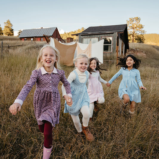 Four young girls running together in a field with rustic cabins in the background. Wearing organic pima cotton childrens dresses with nostalgic styles and playful prints.
